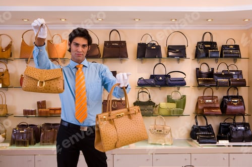 Preview: Front View Portrait Of A Male Shop Assistant In White Gloves Holding Aloft Two Designer Handbag And