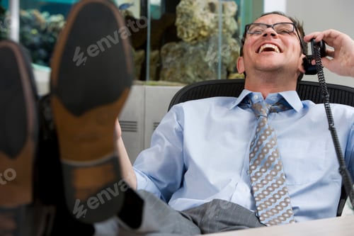 Preview: Horizontal Waist Up Portrait Of A Seated Laughing Businessman With Feet Up On Desk Talking On A