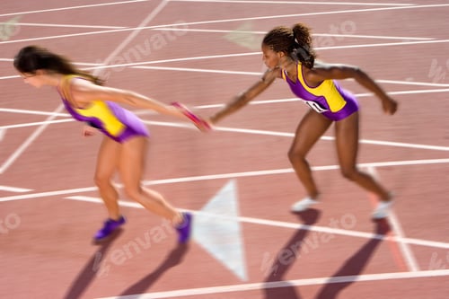 Preview: Wide Angle Shot Of A Young Female Athlete Passing A Relay Baton To Her Teammate On A Bright, Sunny