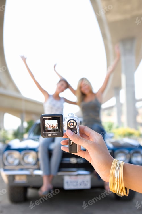 Preview: Close-Up Shot Of A Young Woman'S Hand Taking A Photograph Of Friends On The Bonnet Of A Car Beneath