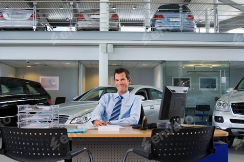Preview: Front View Portrait Of A Smiling Car Salesman Sitting At The Desk In The Large Car Showroom.
