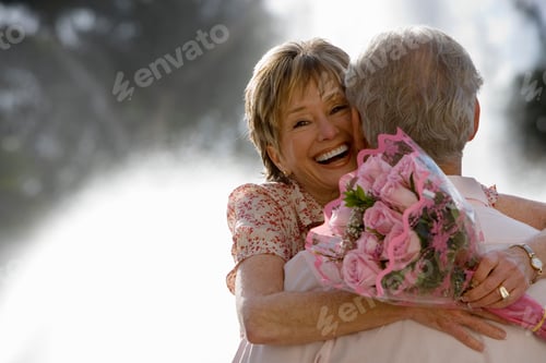 Preview: Portrait Of A Senior Couple Hugging In Front Of A Water Fountain With A Woman Holding A Bouquet Of