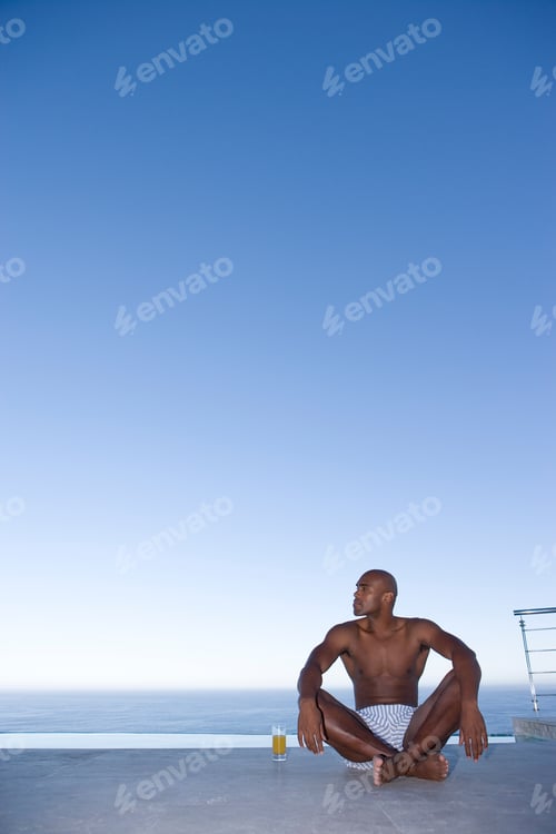 Preview: Young Man Sitting Outdoors On The Ground Next To A Glass Of Juice In Front Of The Sea And Looking