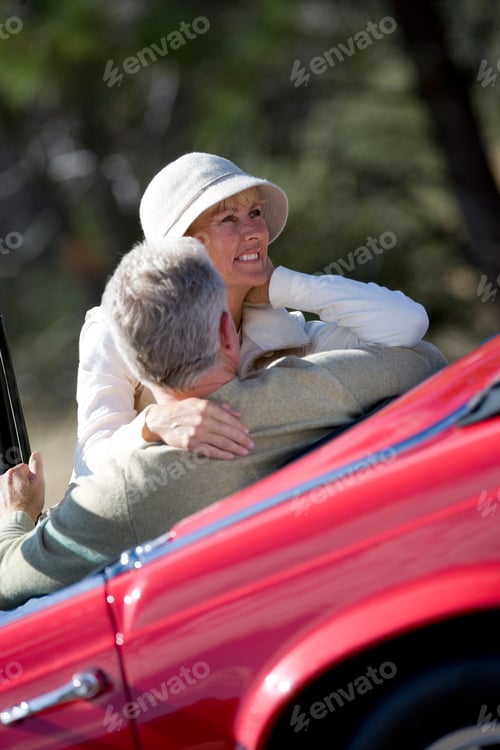 Preview: Tilted Rear View Of A Senior Couple On Road Trip In Red Convertible With Woman Embracing The Man.