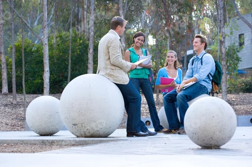 Preview: Full-Length Shot Of Teenage School Students In Conversation With Teacher Sitting On Concrete Balls.