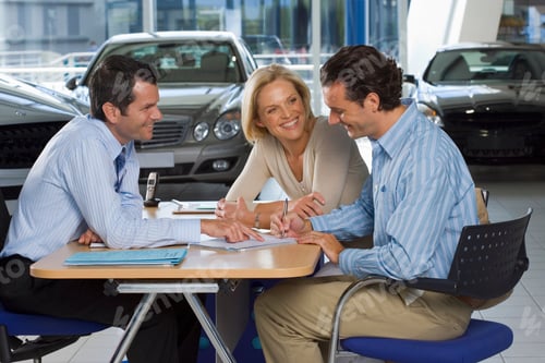 Preview: Side View Of A Car Salesman And A Couple Sitting At The Desk While Man Signs Paperwork In A Large