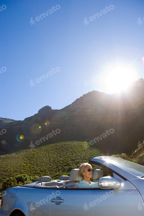 Preview: Vertical Shot Of A Smiling Senior Woman Driving A Convertible Silver Car With Lens Flare Along The
