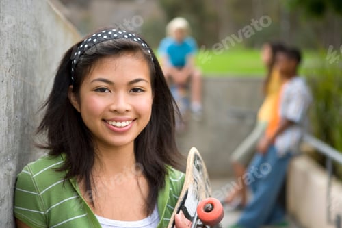Preview: Teenage Girl With Black Hair Holding A Skateboard In Hands And Posing In The Park With Her Friends