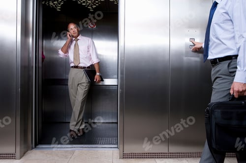 Preview: Businessman On Mobile Phone In Lift With Mid Section Of A Colleague Pressing Button To Call Lift In