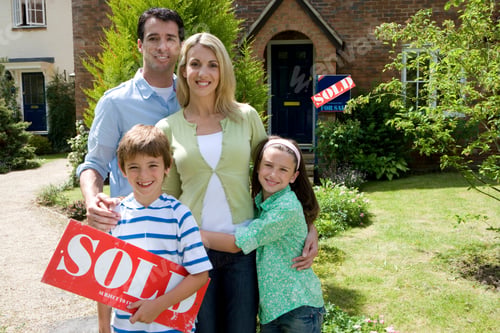Preview: Horizontal Shot Of A Family Of Four Outside A House With Boy Holding A 'For Sale' Sign Smile At The
