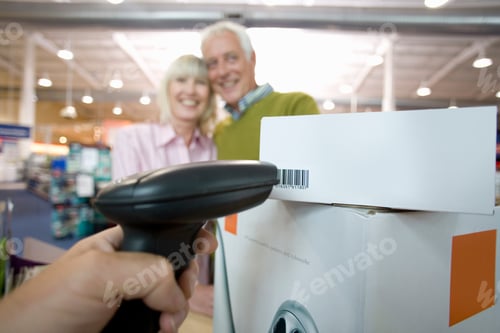 Vista previa: Cajero en el mostrador de facturación escanea un código de barras en una caja mientras una feliz pareja mayor la mira