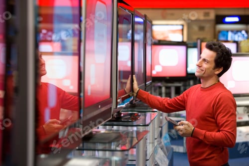 Preview: Young Man Holding A Remote Control In Hand And Standing In The Middle Of An Electronics Appliances