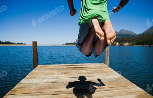 Preview: Horizontal Shot Of Lower Section Of A Boy Jumping Over A Lake Jetty Casting Shadow On A Sunny Day.