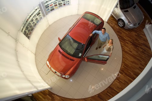 Preview: Elevated Horizontal View Of A Couple Looking At New Red Hatchback In The Car Showroom.