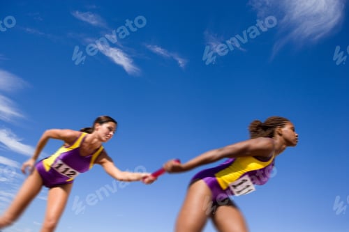 Preview: Young Female Athlete Passing The Relay Baton To Her Teammate On A Bright, Sunny Day At An Athletics