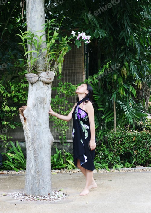 Preview: Asian Woman In A Pretty Summer Sarong Looking At Orchids.