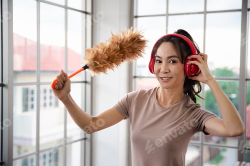 Preview: Young Asian Woman With Headphones Using A Feather Duster To Clean Wood Book Shelves In Home .