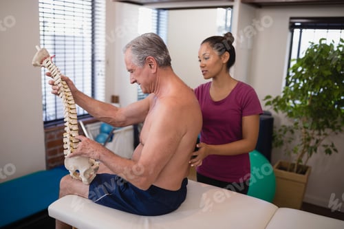 Preview: Shirtless Male Patient Holding Artificial Spine While Female Therapist Examining Back At Hospital