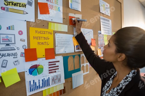 Preview: Female Executive Writing On Sticky Note At Bulletin Board In Office