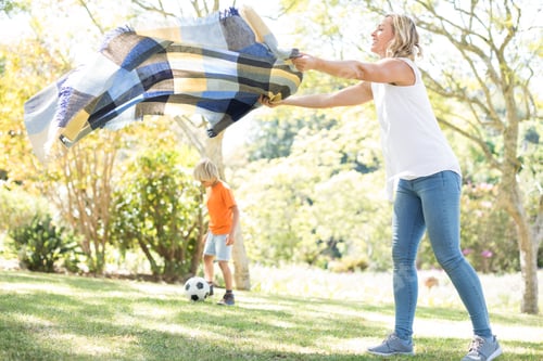 Preview: Mother Spreading The Picnic Blanket While Son Playing Football In Background On A Sunny Day