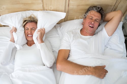 Preview: Annoyed Woman Covering Ears With Pillow While Man Snoring On Bed In Bedroom