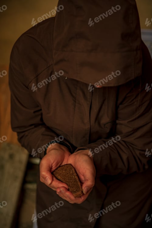 Preview: Girl In Dark Cape Holds Roll And Black Bread In Her Hands. Woman Rehearses Role Of Beggar Woman For