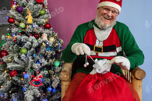 Preview: Elderly Man With Gray Beard And Glasses In Santa Claus Hat With Red Bag Filled With Presents Poses