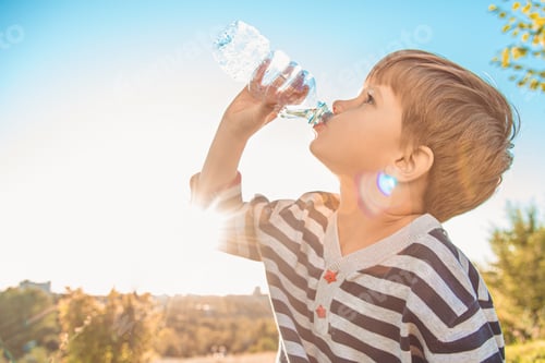 Preview: Cute Boy Sitting On The Grass Drinks Water From A Bottle In The Summer At Sunset. Child Quenches