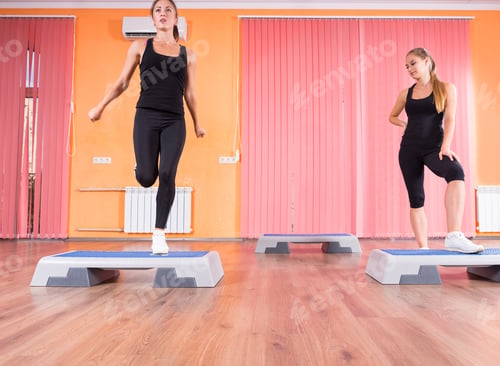 Preview: Low Angle Front View Of Pair Of Young Women Exercising Together In Step Aerobic Class In Colorful