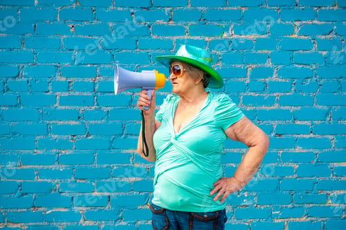 Preview: Elderly Grandmother In Turquoise Sweater And Cowboy Hat Stands With Megaphone Against Gray Brick