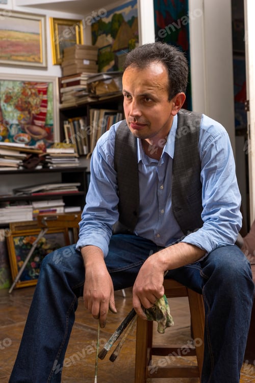 Preview: Male Artist Sitting Thinking In A Studio With A Paint Brush And Rag In His Hand With A Dejected
