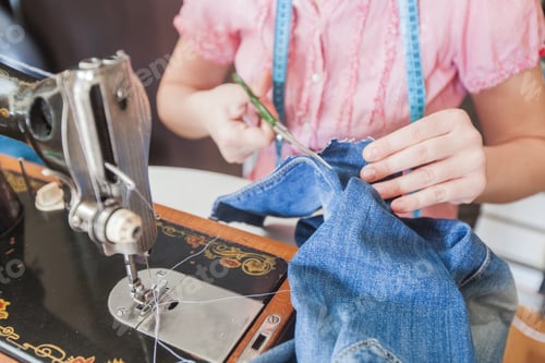 Preview: Female Hands Using Scissors For Cutting Blue Jeans And Retro Sewing Machine At Home.
