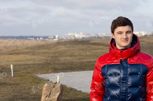 Preview: Handsome Young Man Or Teenager Standing In A Colourful Winter Jacket In The Countryside With A Town