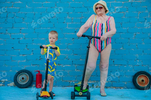 Preview: Elderly Woman In Striped Swimsuit, Sunglasses And Elegant Hat Riding Scooter Against Brick Wall