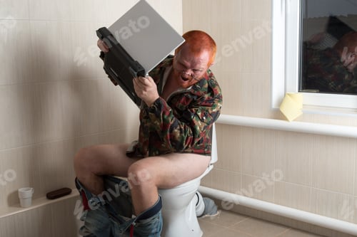 Preview: Man With Red Hair, Beard And Mustache In A Military Jacket In Glasses With Laptop Sitting On Toilet