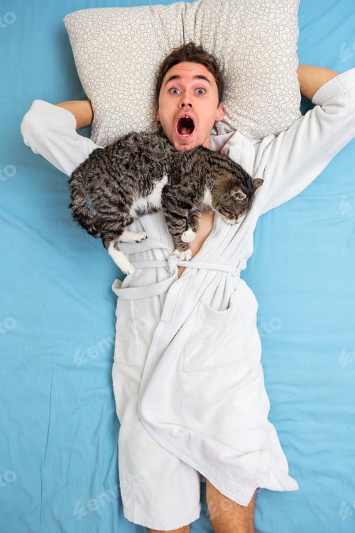 Preview: Top View Of Funny Young Guy In White Bathrobe Lying With Mouth Opened On Bed While Cat Sleeping On