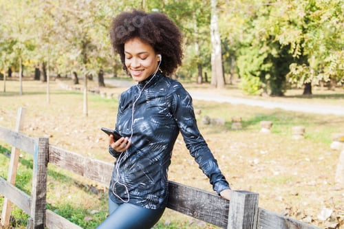 Preview: Sporty African American Woman Relax Listening Music After Training At Park, Leaning On Wooden Fence