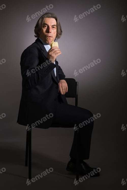 Preview: A Chewing Man With A Pie In His Hand Is Sitting On A Chair On A Black Background.