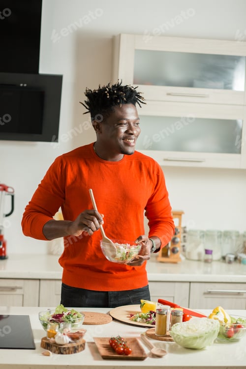 Preview: Positive Young Man Smiling And Looking Into The Distance While Mixing Fresh Salad In The Glass Bowl