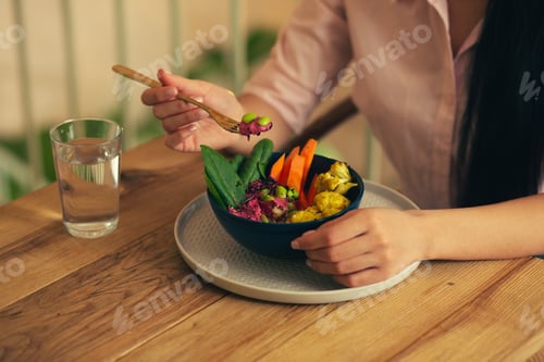 Preview: Woman Sitting At The Wooden Table And Holding A Fork While Eating Her Vegetable Meal From The Bowl