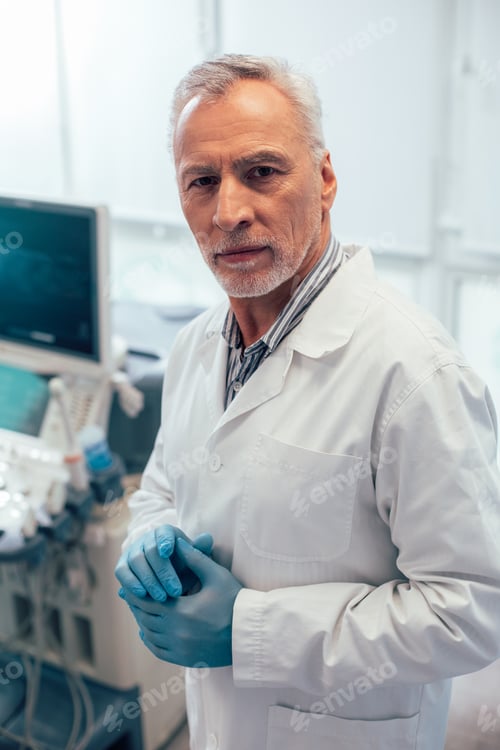 Preview: Calm Mature Male Doctor In A White Coat And Blue Rubber Gloves Standing Alone With Medical