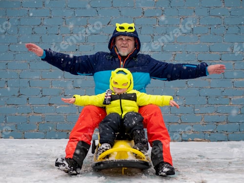 Preview: Dad And Child In Colorful Winter Clothes And Mask Riding Sleigh Along Gray Brick Wall In Snow. Man