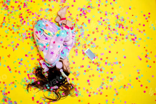 Preview: Happy Little Girl In A Unicorn Kigurumi Lies On A Yellow Background Amid Multicolored Confetti