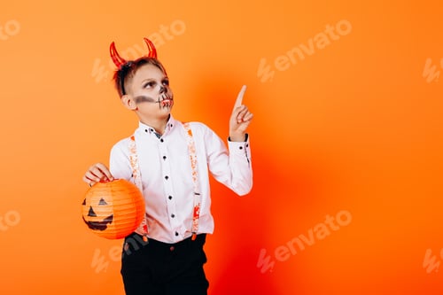 Preview: Boy In Devil Masquerade Makeup Holding A Pumpkin And Pointing Up. Halloween Concept