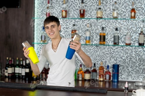 Preview: Happy Young Barman Mixing Cocktails Standing With Colourful Cocktail Shakers In Each Hand With A