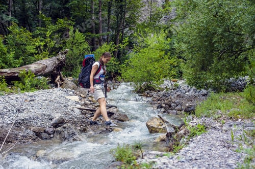 Preview: Caucasian Hiker Woman Crossing The River On Trek In Mountains With Backpack Living A Healthy Active