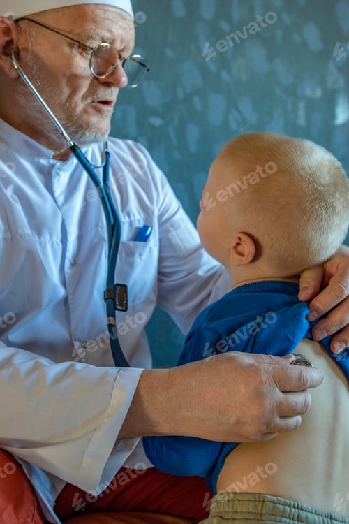 Preview: Home Doctor In White Coat, Cap And Glasses With Gray Beard Examines Two Children Teenagers At Home
