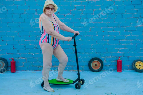 Preview: Elderly Woman In Striped Swimsuit, Sunglasses And Elegant Hat Riding Scooter Against Brick Wall