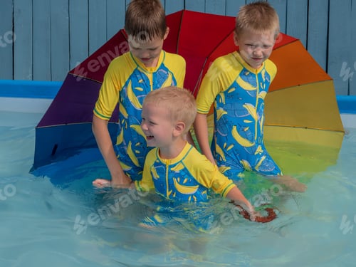Preview: Happy Kids Playing In Carcas Pool In Boys' Swimsuits With Rainbow Umbrella. Brothers Are Happy