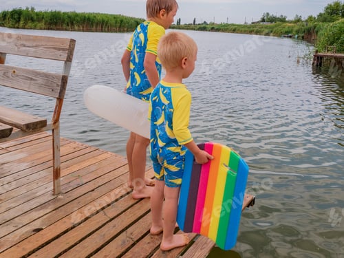Preview: Blond Boy Is Sitting On The Bridge With Multicolor Swimming Board. Child Is Inbathing Suit By